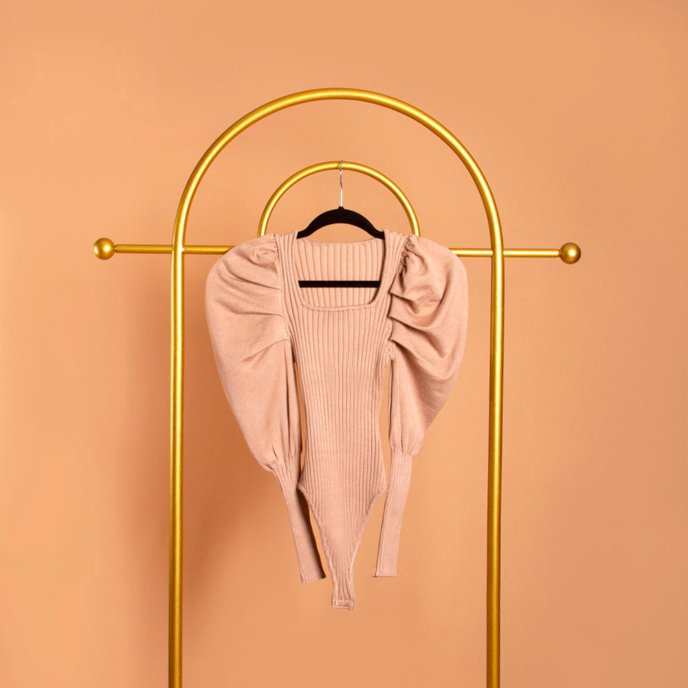 A still image of a tan puff sleeve knit bodysuit on a hanger against an orange wall. 