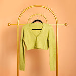 A still image photo of a green rib knit cardigan on a hanger against an orange background.