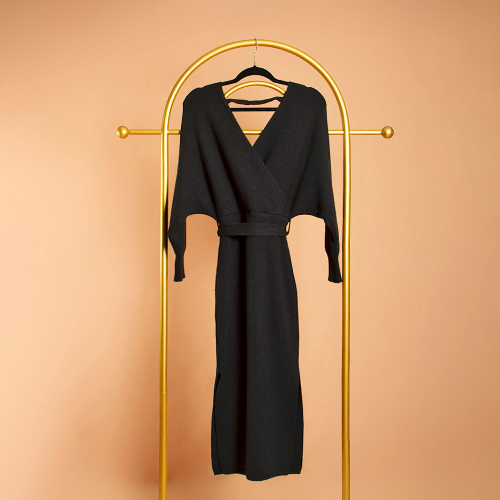 A still image of a black wrap knit midi dress on a hanger against an orange wall. 