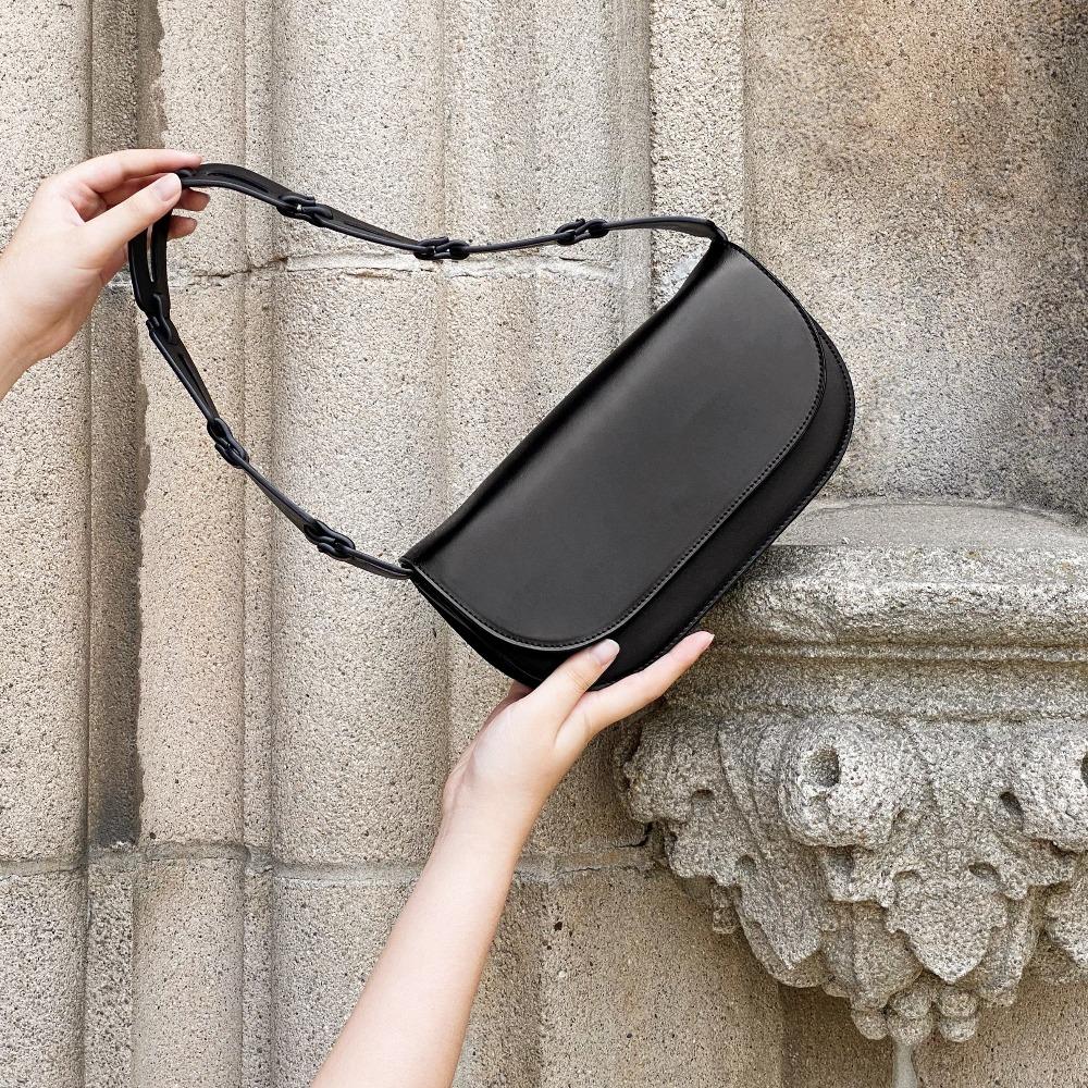 A model holding up a black recycled vegan leather shoulder bag with a scallop strap against a concrete wall. 