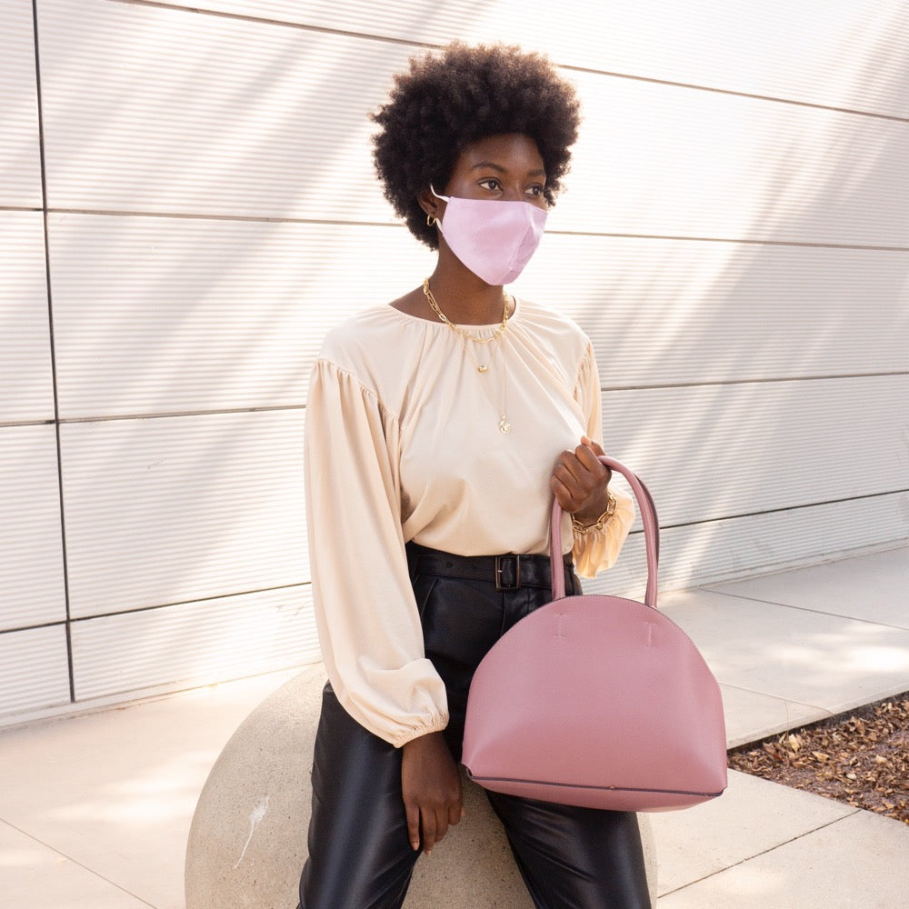 A model wearing a sating lavender face mask with adjustable strings on each side against a white wall outside. 