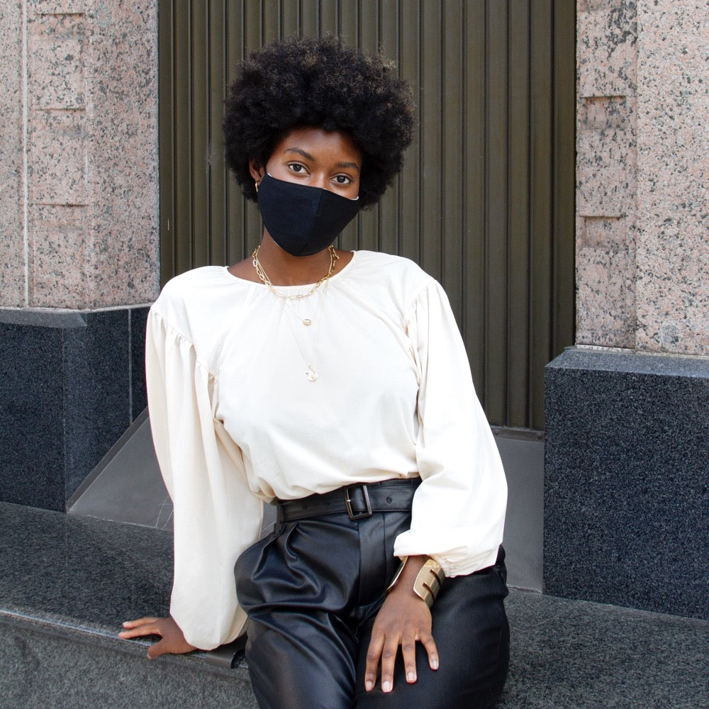 A model wearing a solid black face mask with adjustable string on each side against a wall outside. 