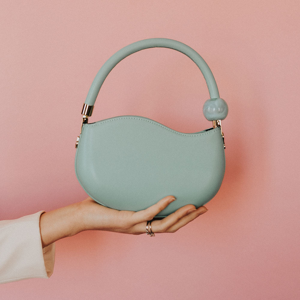 A model holding up a small aqua vegan leather crossbody bag with a marble pearl against a pink wall. 