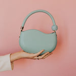 A model holding up a small aqua vegan leather crossbody bag with a marble pearl against a pink wall. 