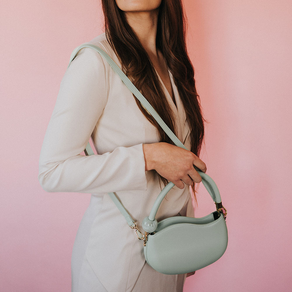 A model wearing a small aqua structured vegan leather crossbody bag against a pink wall. 