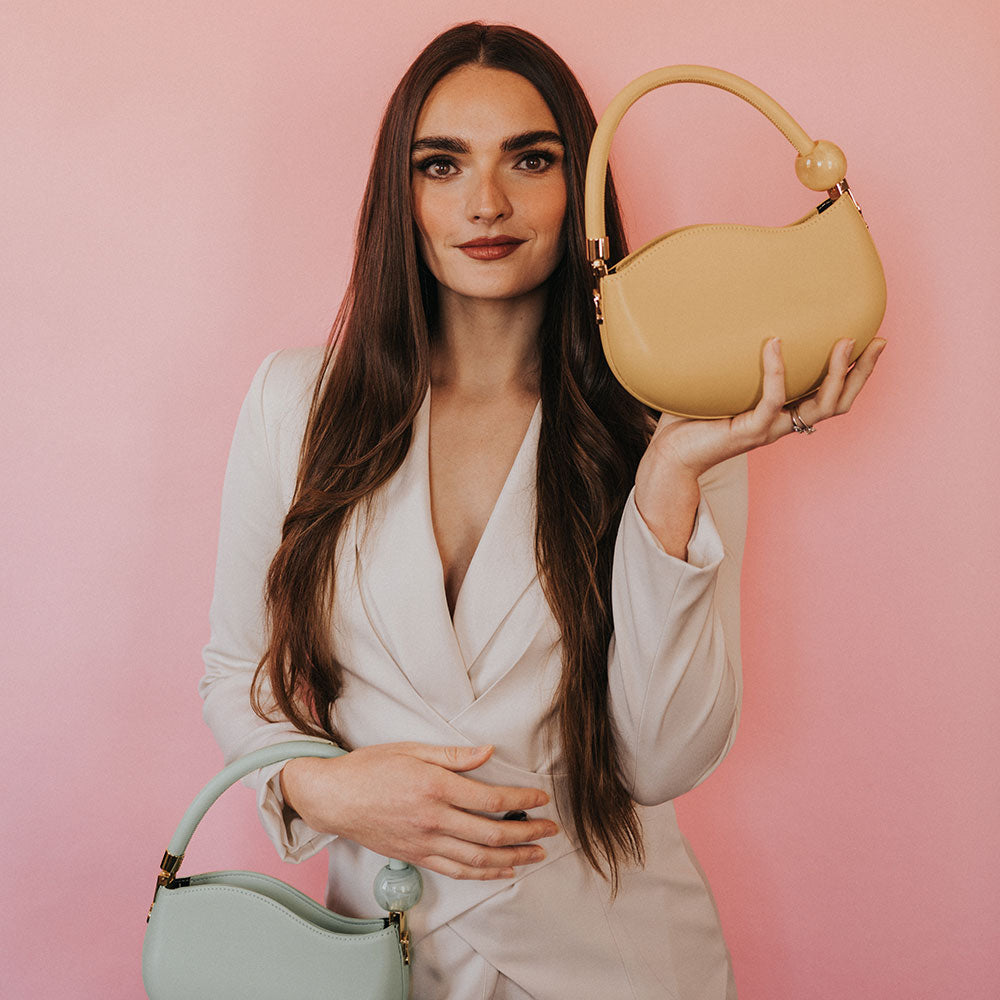 A model holding two structured vegan leather crossbody bags with marble pearl accessory against a pink wall. 