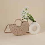 A still image of a small nude semi circle vegan leather top handle bag with pleated detail. Flowers inside a white vase against a tan wall. 