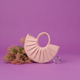 A still image of a small semi circle vegan leather top handle with pleated detail against a purple background with flowers on the side. 