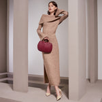 A model wearing a burgundy small vegan leather crossbody bag with pleated detail and wooden handle against a tan wall.