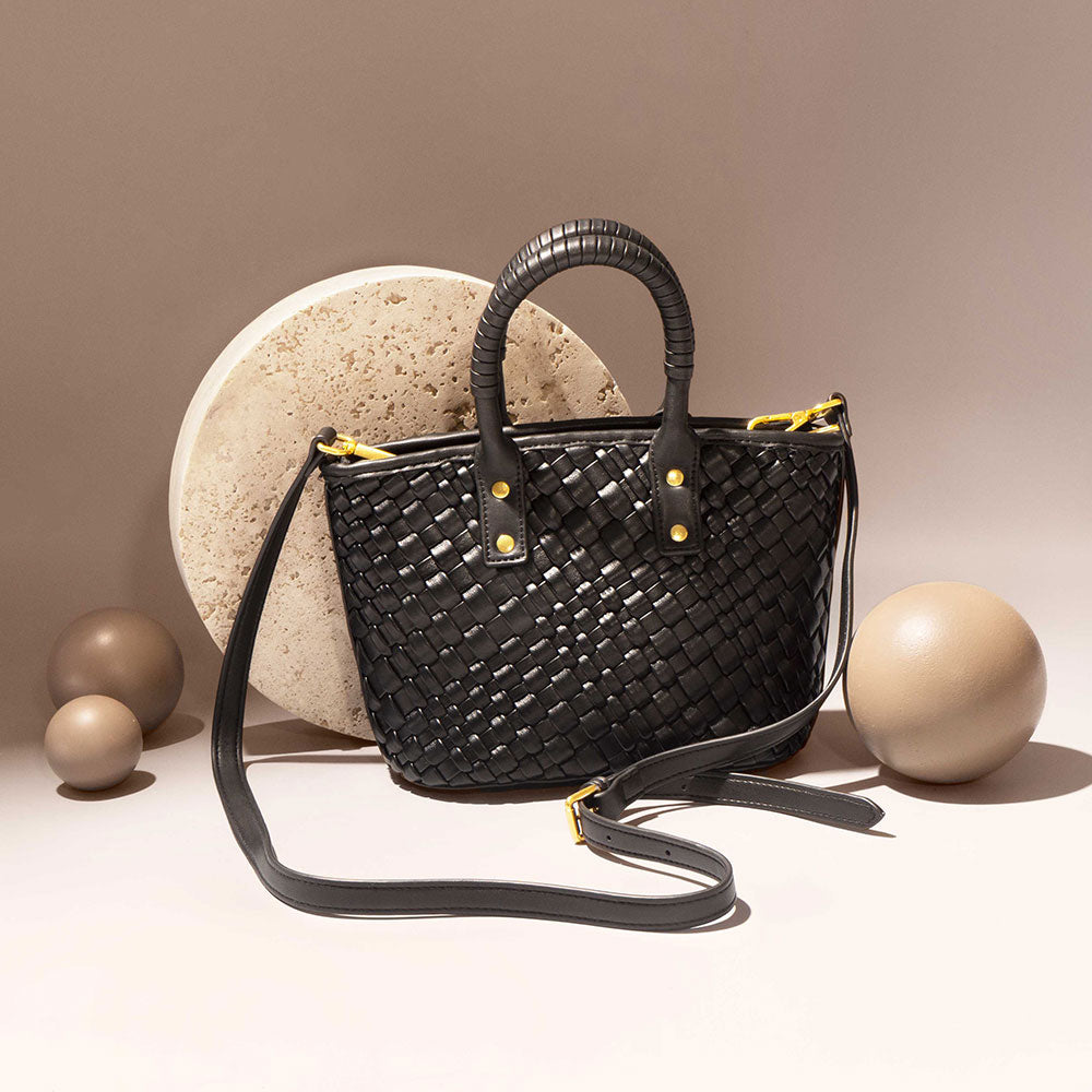 A still image of a woven vegan leather crossbody bag with a wrapped handle against a stone prop. 