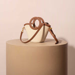 A still image of a canvas and vegan leather crossbody bag with a circle handle against a tan wall. 