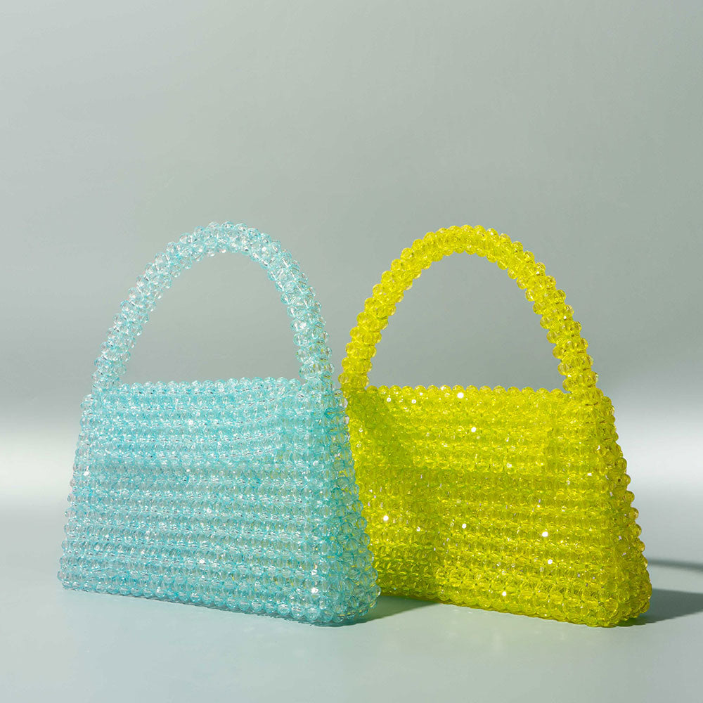 A still image of two crystal beaded top handle bags with a flap closure against a teal wall. 