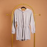 A still image of a grey terry hooded romper on a hanger against a orange wall. 
