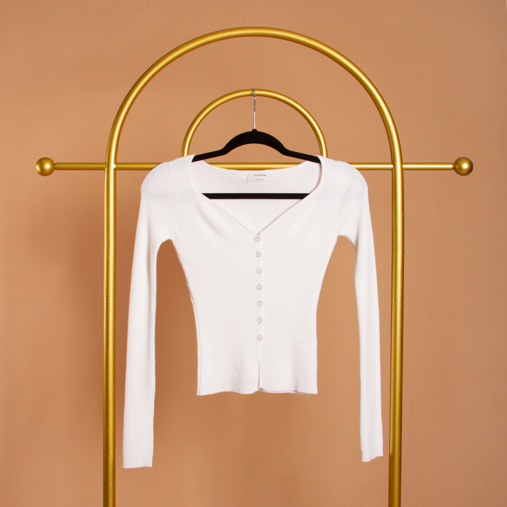 A still image of a white long sleeve rib knit cardigan on a hanger against an orange wall. 