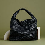 A still image of a large black woven vegan leather shoulder bag against a green wall. 