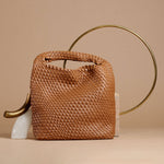 A still image of a large woven vegan leather shoulder bag against a tan wall with gold props.