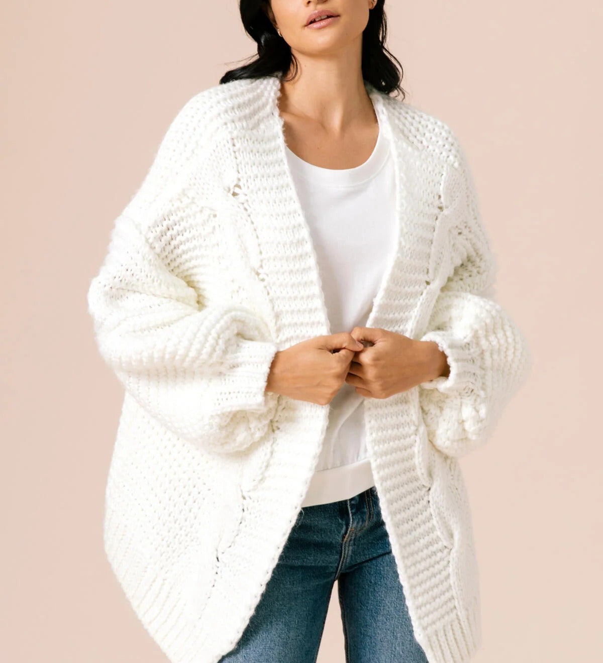 A model wearing a white knitted cardigan against a tan background. 