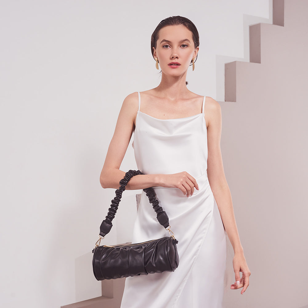 A model wearing a medium cylindrical-shaped shoulder bag with a ruched body and strap against a white wall. 