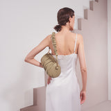 A model wearing a medium moss cylindrical-shaped shoulder bag with a ruched body and strap against a white wall. 