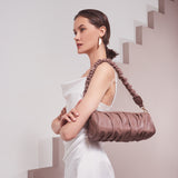 A model wearing a medium taupe cylindrical-shaped shoulder bag with ruched body and strap against a white wall. 