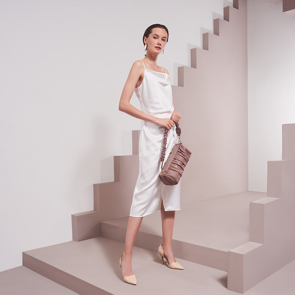 A model wearing a medium taupe cylindrical-shaped shoulder bag with ruched body and strap against a white wall. 
