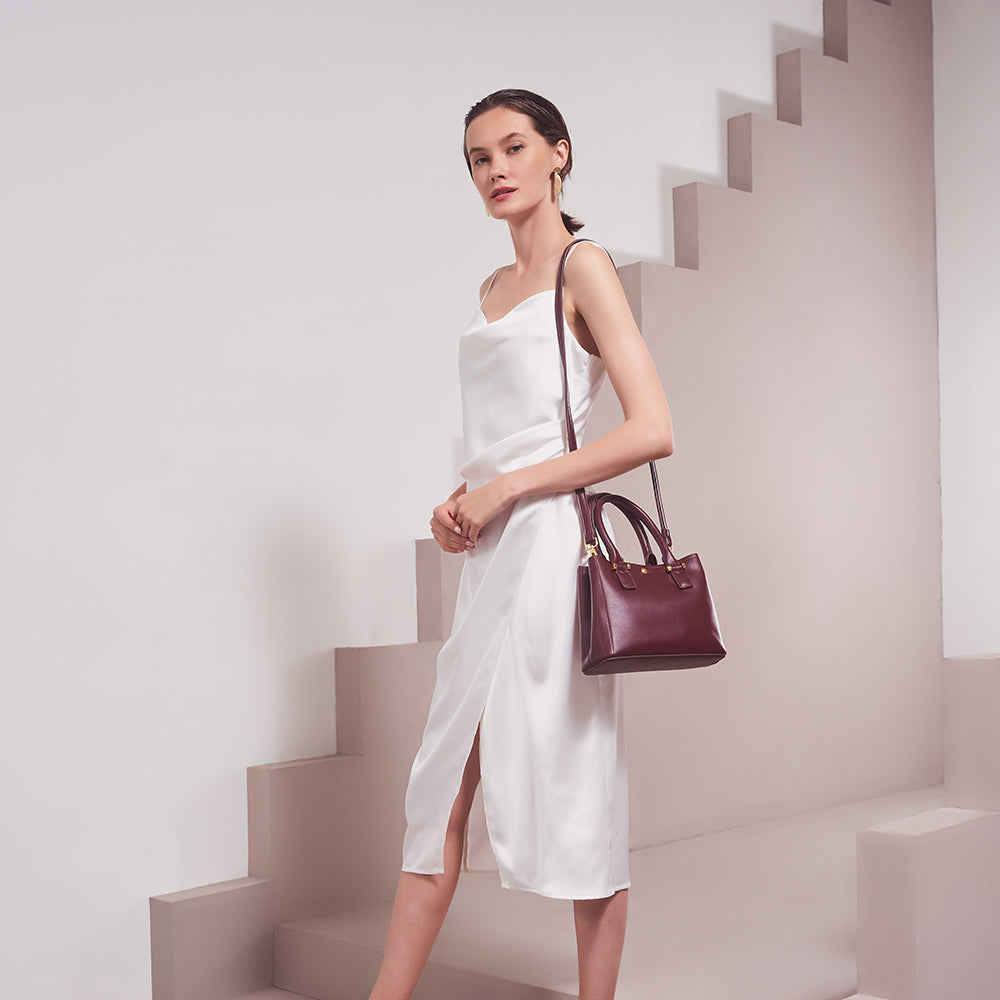 A model wearing a small burgundy recycled vegan leather crossbody handbag against a tan background. 