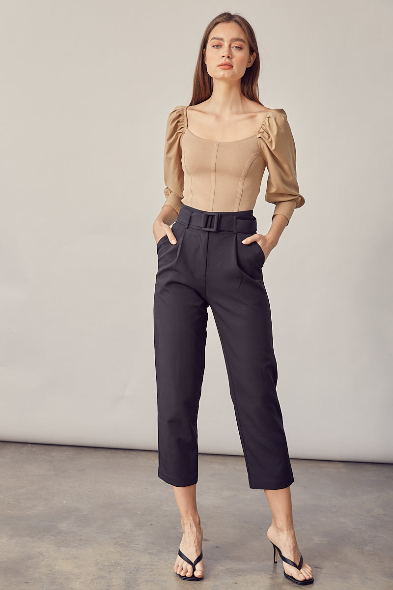 A model wearing a black straight leg pant with a belt against a white wall. 