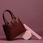 A still image of a small burgundy crossbody handbag against a pink wall with pink props. 