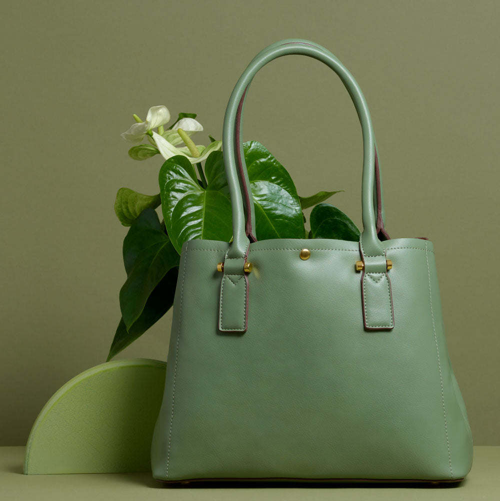 A still image of a large sage vegan leather shoulder bag with flowers inside against a green wall. 