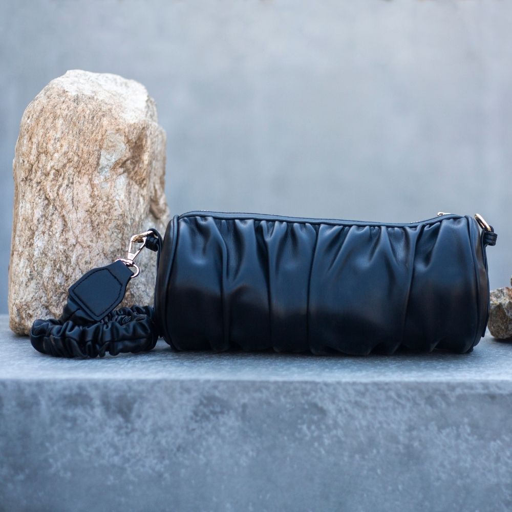 A still image of a medium black cylindrical-shaped shoulder bag with a ruched body and strap sitting on a rock.