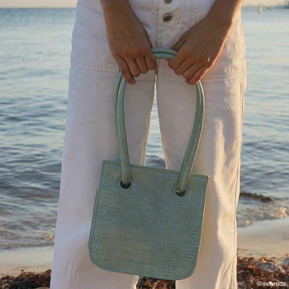 A model holding a medium shiny croc shoulder bag with water in the background.