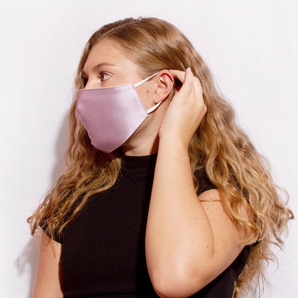 A model wearing a lavender satin face mask with adjustable strings on each side against a white wall. 